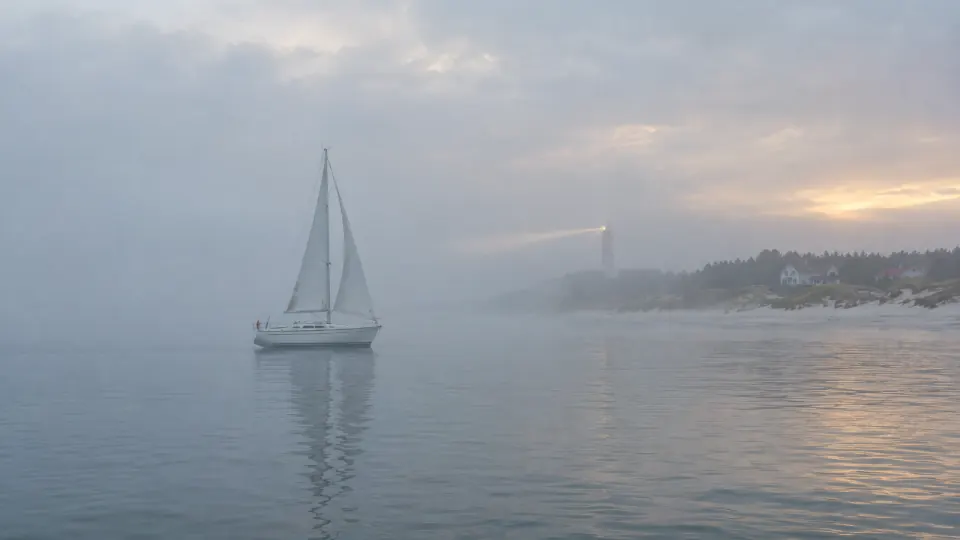 Weiße Segelyacht im dichten Morgennebel, in der Ferne leuchtet schwach ein Leuchtturm an der Küste