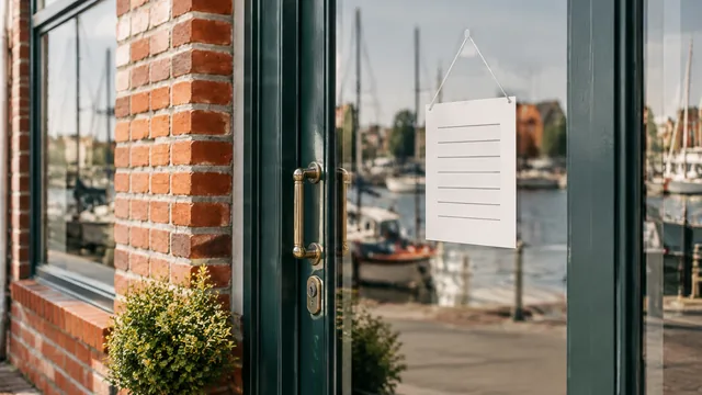 Eine Ladentür aus Glas mit Messinggriff, an der ein leeres Schild für Öffnungszeiten hängt, in der Scheibe spiegelt sich ein Hafen mit Segelbooten