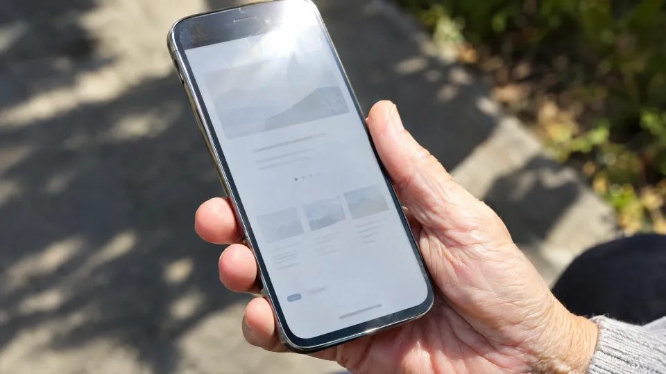 Ältere Hand hält ein Smartphone in der Sonne, auf dem Bildschirm eine Website mit blassgrauer, kaum lesbarer Schrift