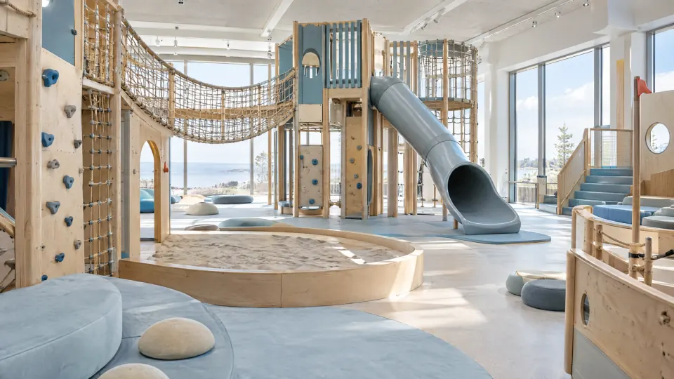 Heller Indoor-Spielbereich mit Kletterturm aus Holz, Netzbrücke, Rutsche und Sandkasten vor großen Fenstern