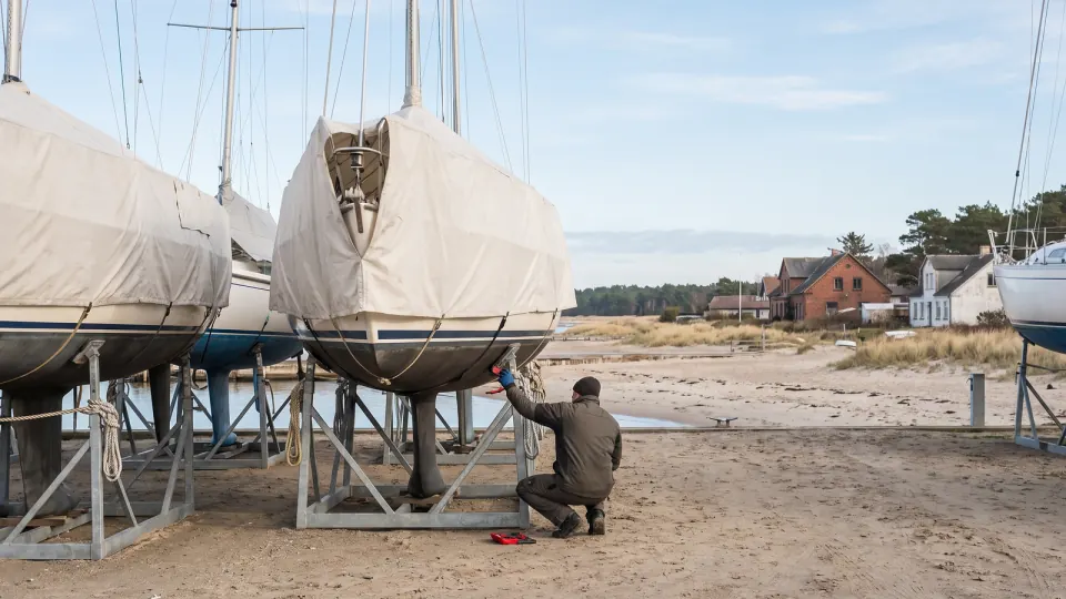 Segelboote im Winterlager an Land, eine Person prüft einen Rumpf
