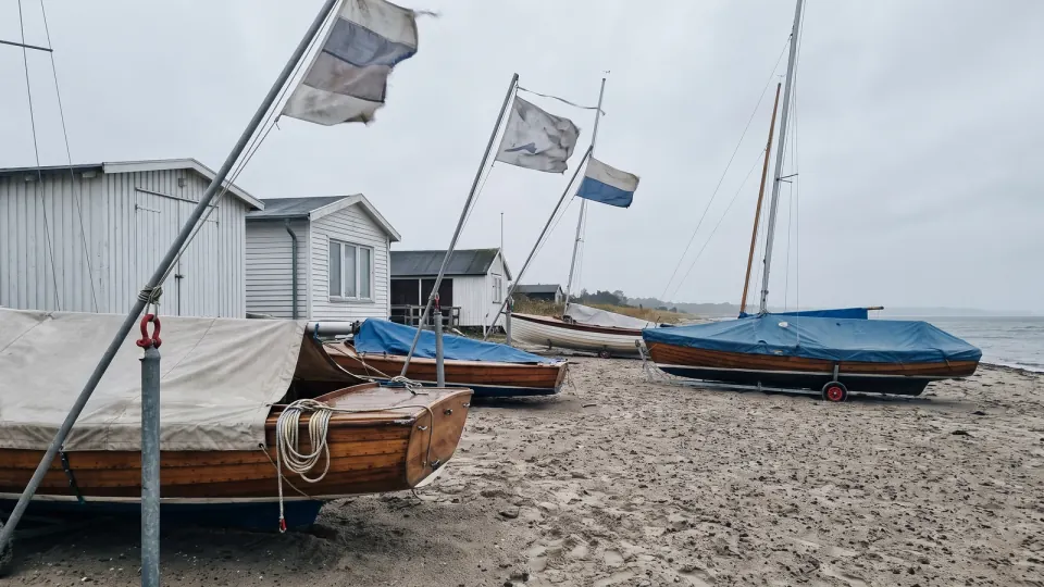 Abgedeckte Jollen am Strand vor Bootsschuppen an einem windigen, grauen Tag