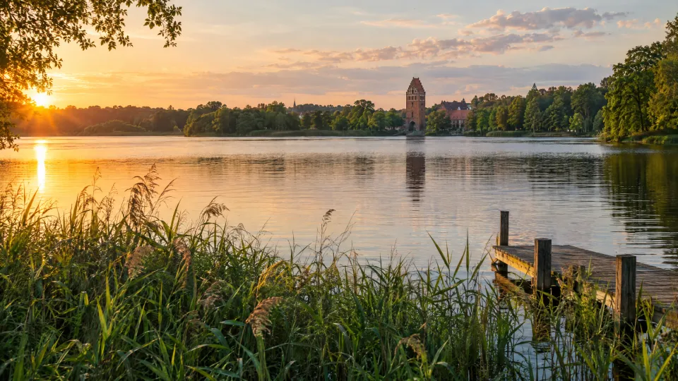 Symbolbild, KI-generiert: See mit Schilf und Holzsteg im Abendlicht, am anderen Ufer ein Backsteinturm, angelehnt an den Tollensesee