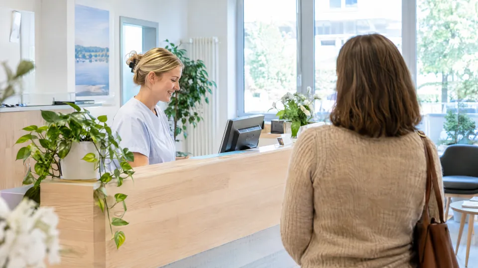 Heller Empfangsbereich einer Hausarztpraxis mit Holztresen, eine Medizinische Fachangestellte und ein wartender Patient, viel Tageslicht