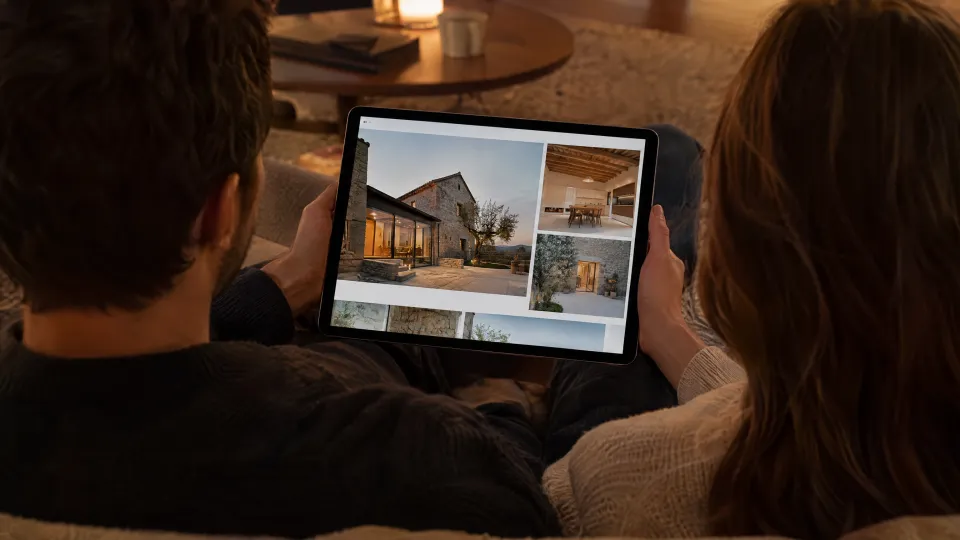 Paar von hinten auf dem Sofa mit einem Tablet, auf dem ein Architektur-Portfolio mit großen Fotos eines sanierten Hauses zu sehen ist