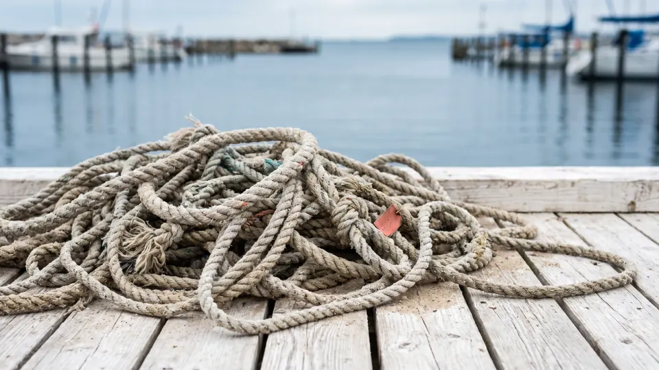 Verhedderte alte Festmacherleinen auf einem Holzsteg, dahinter ein ruhiger Hafen